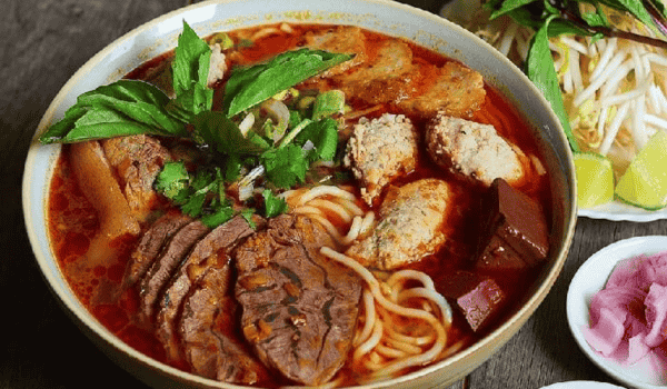 Un bol de bún bò Huế coloré, typique de la gastronomie vietnamienne d'été, garni de bœuf, pâté de crabe, boudin, herbes fraîches et nouilles dans un bouillon épicé.