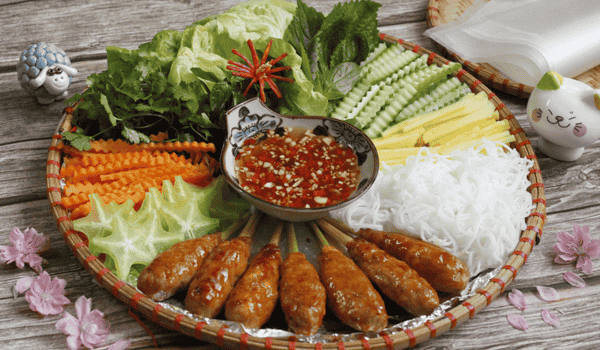 Des brochettes de nem lụi grillées sur charbon, servies avec des galettes de riz, des herbes fraîches, des légumes croquants et une sauce aux arachides – une spécialité incontournable de la gastronomie vietnamienne d'été.